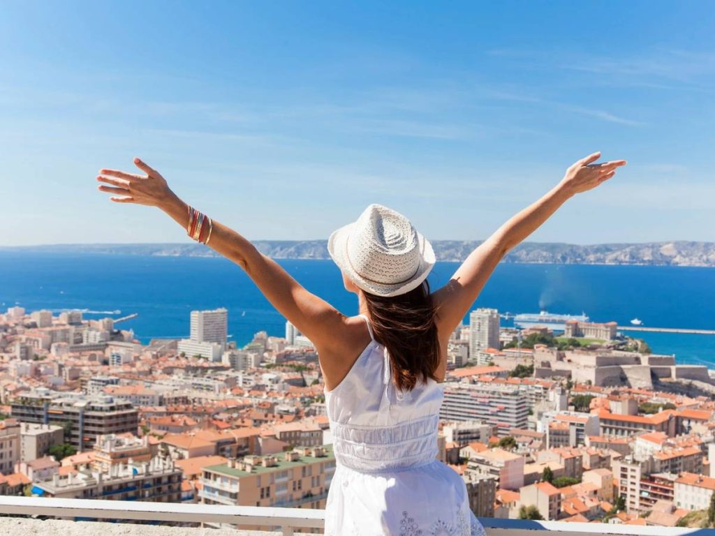 girl flinging her arms wide overlooking a blue sea and city loves practical travel
